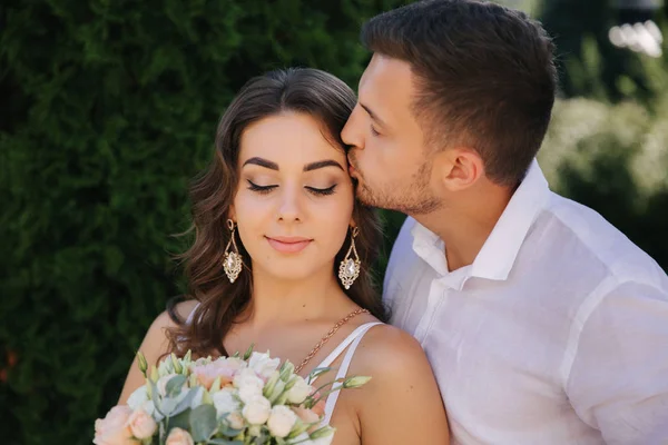 Um homem barbudo e bonito beija a mulher. Mulher gogeous em vestido branco com belo buquê. Homem elegante em camisa branca — Fotografia de Stock