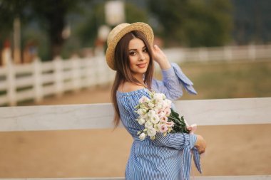Georgeus kadın çiftlik önünde stand ve bir buket çiçek elinde tut. Beautuful buket. Kadın mavi elbise ve örme şapka