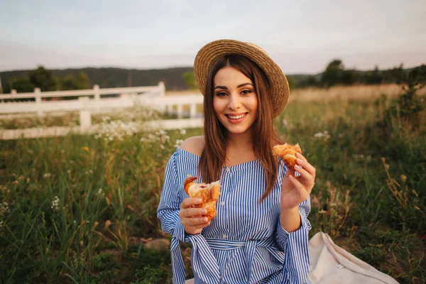 Mujer embarazada en el campo comiendo croissant crujiente respaldado fresco. Comida saludable. La joven come muy apetitoso. Contexto de la explotación — Foto de Stock