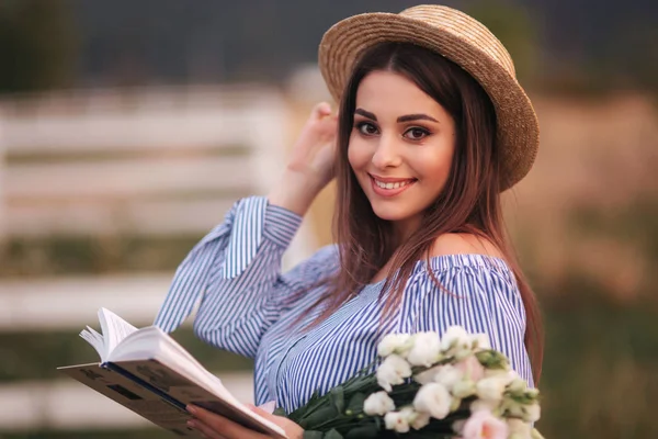 Елегантна жінка тримає в руці книгу і букет квітів. Вона стоїть у полі. Фермерське господарство. Читання книги про дитину — стокове фото