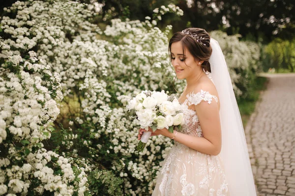 Noiva encantadora em lindo vestido de noiva segurar buquê e ficar na frente de árvores florescendo — Fotografia de Stock