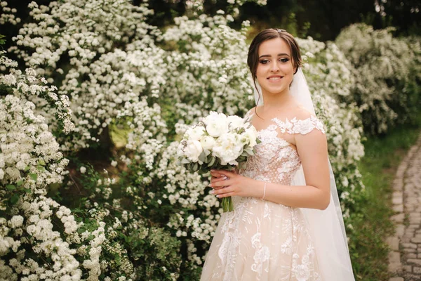 Noiva encantadora em lindo vestido de noiva segurar buquê e ficar na frente de árvores florescendo — Fotografia de Stock