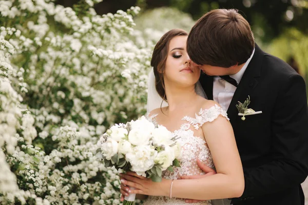 Knappe bruidegom knuffel zijn bruid in hun trouwdag. Achtergrond van bloeiende bomen — Stockfoto