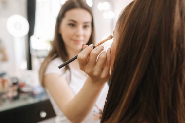 Güzel femalemakeup sanatçı büyük bir ayna önünde oturan bir güzellik salonunda genç bir Redhead kız için makyaj yapıyor. Tatil ve toplantı için hazırlık konsepti
