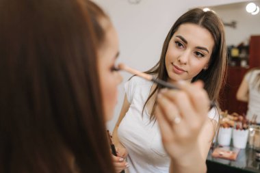 Güzel femalemakeup sanatçı büyük bir ayna önünde oturan bir güzellik salonunda genç bir Redhead kız için makyaj yapıyor. Tatil ve toplantı için hazırlık konsepti