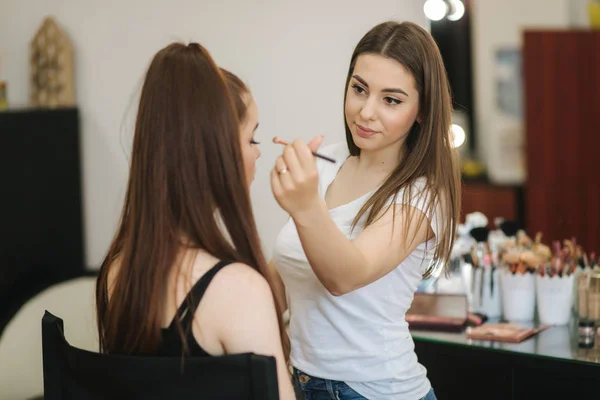 Schöne Femalemakeup-Artist Make-up für ein junges rothaariges Mädchen in einem Schönheitssalon sitzt vor einem großen Spiegel. Konzept der Vorbereitung auf den Urlaub und das Treffen — Stockfoto