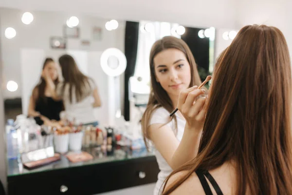 Linda artista femalemakeup fazendo maquiagem para uma jovem ruiva em um salão de beleza sentado na frente de um grande espelho. Conceito de preparação para o feriado e reunião — Fotografia de Stock