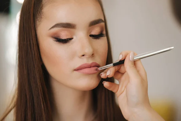 Makeup artysta stosuje szminkę. Ręka makijażu mistrza, malowanie usta młodego modelu rude piękno. Makijaż w procesie. Widok zbliżenie — Zdjęcie stockowe