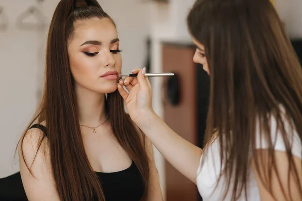 Bella truccatrice applica il rossetto. Mano di maestro di trucco, dipingendo labbra di giovane bellezza modello rossa. Make up in processo . — Foto Stock