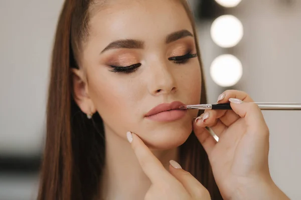 Maquillaje artista aplica lápiz labial. Mano de maquillador maestro, pintando labios de joven modelo pelirroja belleza. Maquillaje en proceso. Vista de cerca — Foto de Stock