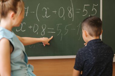 Genç bayan öğretmen okul çocuğuna yardım ediyor. Çocuk tahtanın yanında dur.