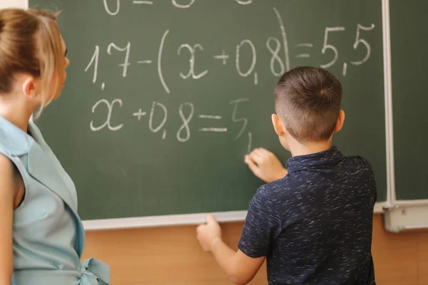 Una maestra joven ayuda a un escolar. Chico de pie junto a la pizarra — Foto de Stock