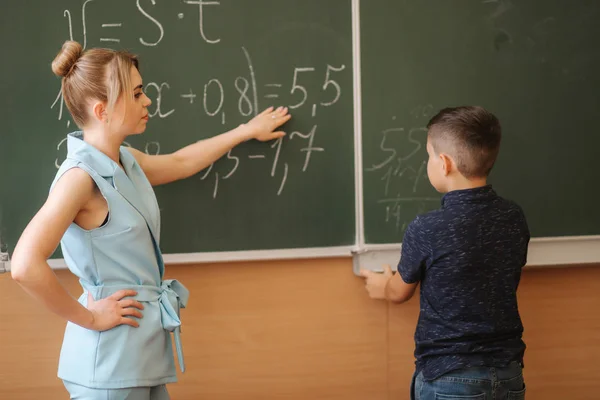 Νεαρή δασκάλα βοηθάει το σχολιαρόπαιδο. Αγόρι στάσου δίπλα στον πίνακα. — Φωτογραφία Αρχείου