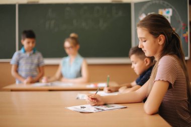 Eğitim, ilkokul. Öğrenme ve insan konsepti - kalemi ve not defterleri olan bir grup okul çocuğu sınıfta yazı sınavı