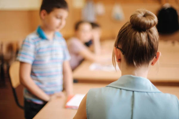 Professor bonito na sala de aula sentado na mesa e pedindo crianças. educação, escola primária, aprendizagem e conceito de pessoas — Fotografia de Stock