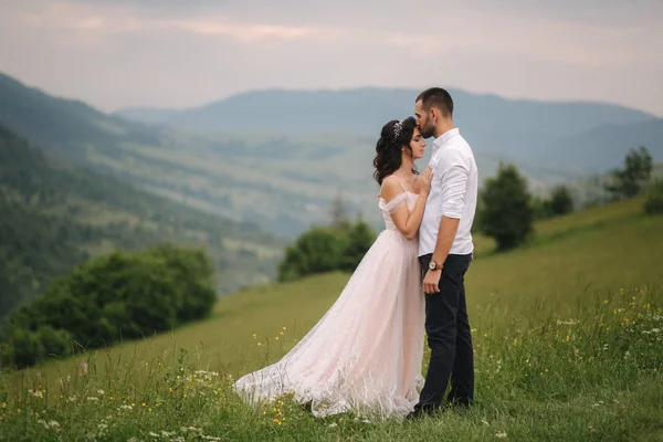 Bella giovane coppia di nozze in piedi sul pendio verde, collina. Sposo e sposa sulle montagne dei Carpazi. Uomo bacio donna in fronte — Foto Stock