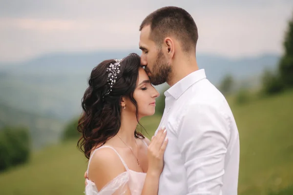 Casal jovem bonito de pé na encosta verde, colina. Noiva e noivo nas montanhas dos Cárpatos. Homem beijo mulher na testa — Fotografia de Stock