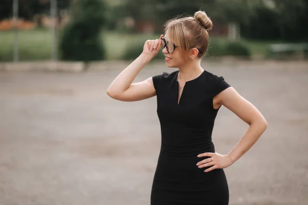 Jonge aantrekkelijke vrouwelijke techer in zwarte jurk met zonnebril buiten staan. Achtergrond van school — Stockfoto