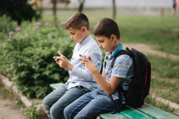 Två pojke sitter på bänken och spela online-spel. En pojke med ryggsäck. Unga pojkar använder sina telefoner — Stockfoto