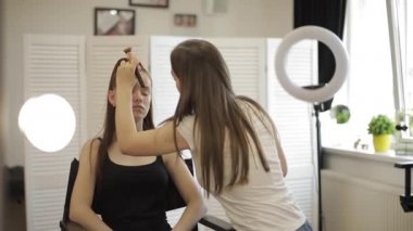 Güzel kadın makyaj sanatçısı büyük bir ayna önünde oturan bir güzellik salonunda genç bir Redhead kız için makyaj yapıyor. Tatil ve toplantı için hazırlık konsepti