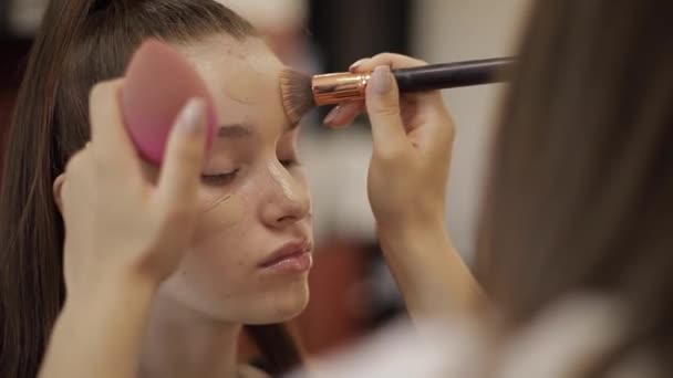 Linda artista de maquiagem feminina fazendo maquiagem para uma jovem ruiva em um salão de beleza sentado na frente de um grande espelho. Conceito de preparação para o feriado e reunião — Vídeo de Stock