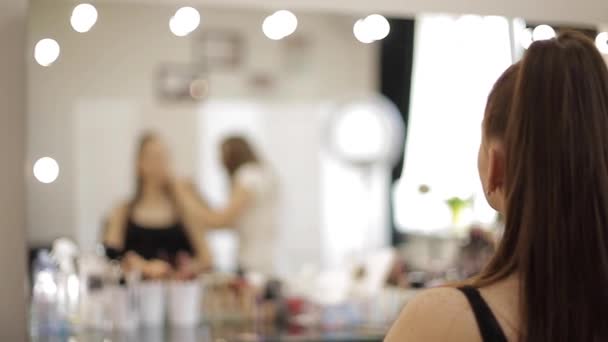 Красива художниця-візажистка робить макіяж для молодої рудої дівчини в салоні краси, що сидить перед великим дзеркалом. Концепція підготовки до свята та зустрічі — стокове відео