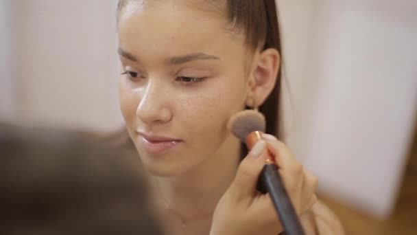 Beautiful female makeup artist doing makeup for a young redhead girl in a beauty salon sitting in front of a large mirror. Concept of preparation for the holiday and meeting — Stock Video