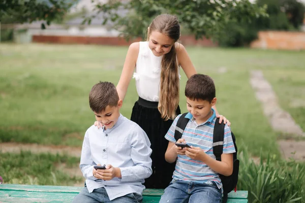 Dva kluci a holka používají telefony během školních přestávek. Roztomilí kluci sedí na lavičce a hrát online hry — Stock fotografie