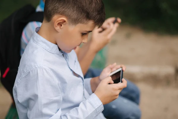 Två pojke sitter på bänken och spela online-spel. En pojke med ryggsäck. Unga pojkar använder sina telefoner — Stockfoto