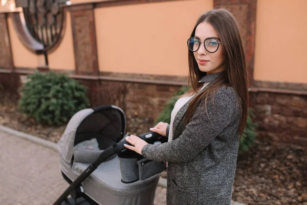 Genç Anne, Bebek arabası içinde yenidoğan bebeği ile yürüyor. Gri bir Pram. Güzel anne içinde Sunglasses