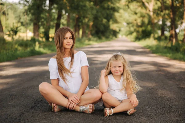 Attraktive Mutter und blonde Haare Tochter sitzt auf der Straße in der Nähe der großen Gasse. sie lächeln und schauen zur Natur. Frontansicht — Stockfoto