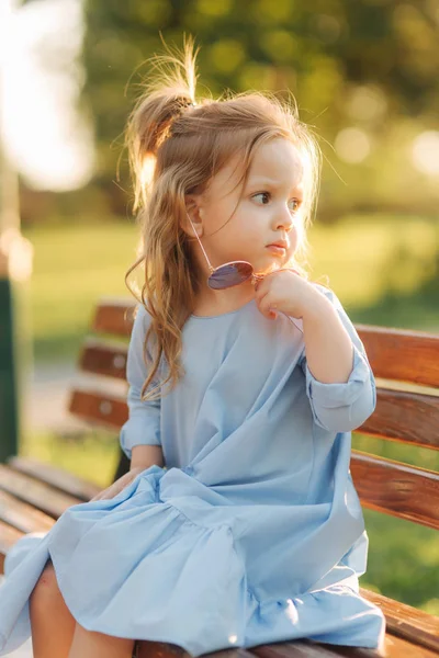 Μικρό κορίτσι μοντέλο σε μπλε φόρεμα και γυαλιά ηλίου που κάθεται σε ένα παγκάκι στο πάρκο — Φωτογραφία Αρχείου