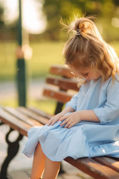 Modelo niña en vestido azul y gafas de sol se sienta en un banco en el parque — Foto de Stock