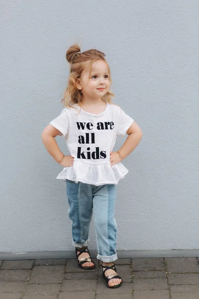 Schöne kleine Mädchen in weißem Hemd und Jeans stehen vor grauer Wand. Glückliche Kinder — Stockfoto