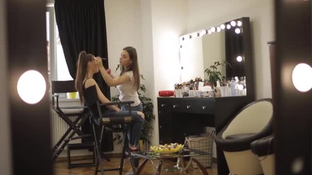 Güzel kadın makyaj sanatçısı büyük bir ayna önünde oturan bir güzellik salonunda genç bir Redhead kız için makyaj yapıyor. Tatil ve toplantı için hazırlık konsepti — Stok video
