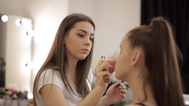 Beautiful female makeup artist doing makeup for a young redhead girl in a beauty salon sitting in front of a large mirror. Concept of preparation for the holiday and meeting — Stock Video