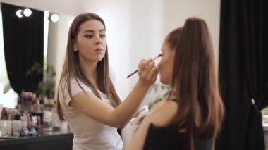 Güzel femalemakeup sanatçı büyük bir ayna önünde oturan bir güzellik salonunda genç bir Redhead kız için makyaj yapıyor. Tatil ve toplantı için hazırlık konsepti