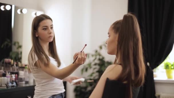 Hermosa femalemakeup artista haciendo maquillaje para una joven pelirroja en un salón de belleza sentado frente a un gran espejo. Concepto de preparación para las vacaciones y la reunión — Vídeo de stock