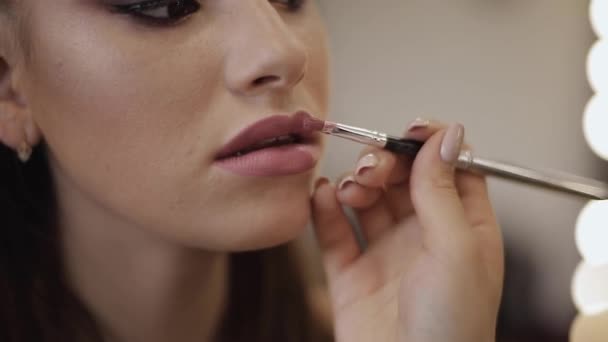 Make-up aplikuje rtěnku. Ruka make-up mistra, malířské rty mladého modelu rudhead krásy. Doplní se do procesu. Zobrazení closeup. Video — Stock video