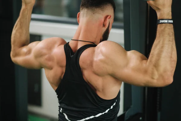 Un joven atleta entrena en el gimnasio. Muestra los músculos de la espalda y el pecho. Vista trasera — Foto de Stock