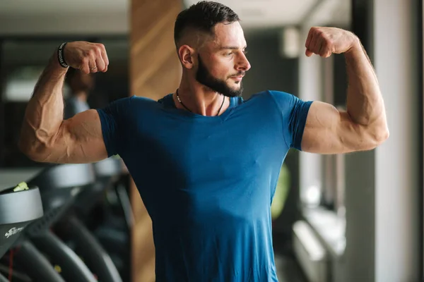 Um jovem atleta de t-shirt azul treina no ginásio. Mostra os músculos — Fotografia de Stock