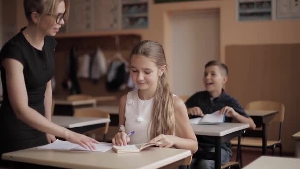 Lärare hjälper skolbarn att skriva prov i klassrummet. utbildning, grundskola, lärande och människor koncept — Stockvideo