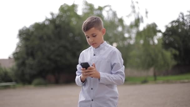 Το όμορφο αγόρι χρησιμοποιεί τηλέφωνο έξω. Το αγοράκι στην πίσω αυλή του σχολείου. — Αρχείο Βίντεο