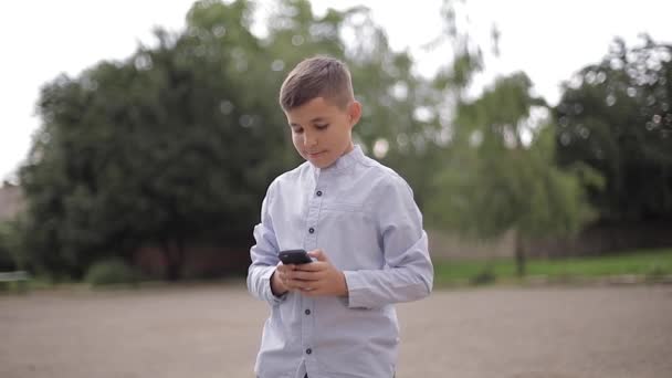 Pohledný chlapec používá telefon venku. Malý chlapec na zadním dvoře školy — Stock video