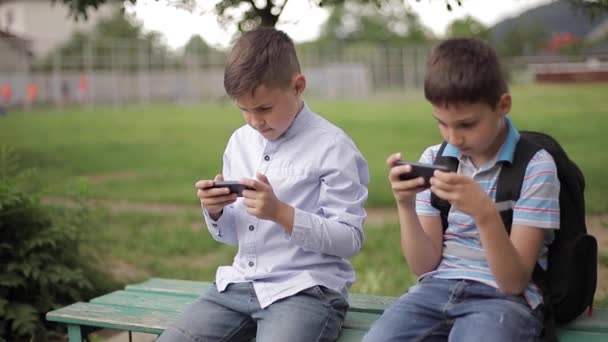 Dwa chłopiec siedzi na ławce i grać w gry online. Jeden chłopcy z plecakiem. Młodzi chłopcy używają swoich telefonów — Wideo stockowe