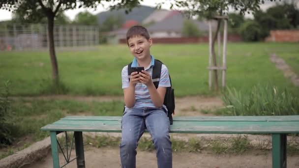 Giovane ragazzo con lo zaino seduto sulla panchina e giocare online durante le vacanze scolastiche — Video Stock