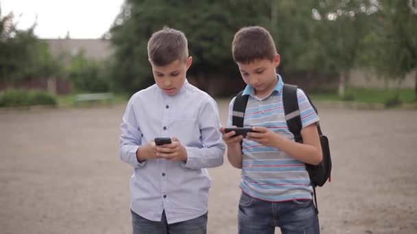 Два мальчика играют в онлайн-игры после уроков. Мальчики улыбаются и пользуются телефоном — стоковое видео