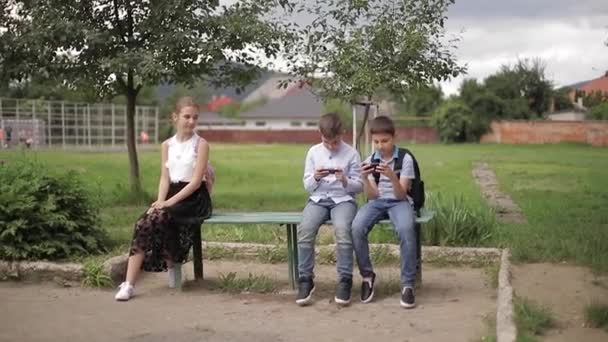 Deux garçons et une fille utilisent leur téléphone pendant la pause scolaire. Garçons mignons assis sur le banc et jouer à des jeux en ligne — Video