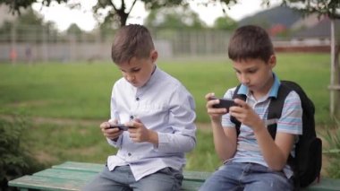 İki çocuk bankta oturuyor ve online oyunlar oynuyor. Sırt çantası olan bir çocuk. Genç çocuklar kendi telefonları kullanın
