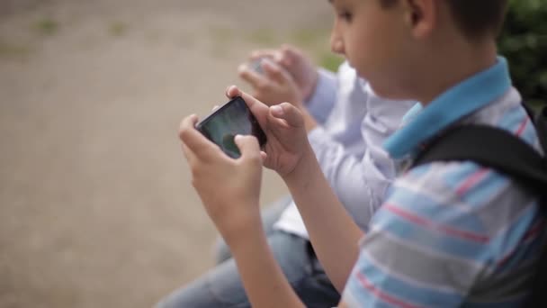 İki çocuk bankta oturuyor ve online oyunlar oynuyor. Sırt çantası olan bir çocuk. Genç çocuklar kendi telefonları kullanın — Stok video